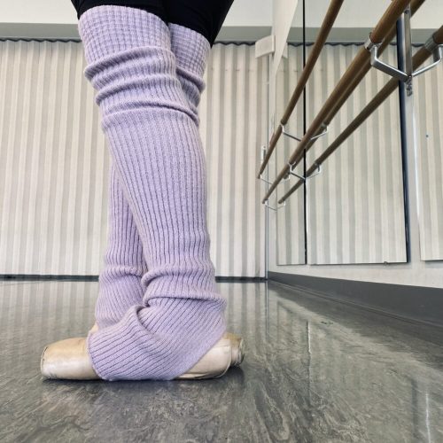 A person wearing leg warmers and pointe shoes standing in a ballet 5th position at a ballet barre.